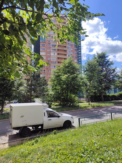 In Putilkovo, Russia, a white van with a cargo box is parked on the side of a street. The van is positioned at an angle, facing towards the right of the frame, with its rear left wheel visible. The street is paved, and a green metal fence runs alongside it. Beyond the fence, a lush green lawn slopes upwards. In the background, a tall, multi-story residential building with a brick facade and numerous windows stands out against a bright blue sky dotted with white clouds. Several trees with green foliage are scattered in front of the building and along the street, with some branches of a leafy tree in the foreground partially obscuring the view.