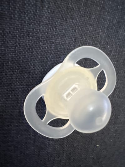 A pacifier rests on a dark, textured surface, likely fabric. The pacifier is translucent white and features a rounded nipple and a shield with two oval-shaped openings. A small, raised marking is visible on the shield, possibly a brand logo. The lighting appears to be overhead, creating subtle shadows. The image does not provide any information about the city of Moelv, Norway, or any other location.