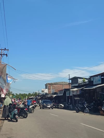 In the city of Deah Pangwa, Indonesia, a street scene unfolds under a clear blue sky with a few wispy clouds. The road is paved with asphalt and marked with white lines. On the right side of the road, a row of shops with corrugated metal roofs are visible, some displaying signs, including one that reads "PUTRA DAYAH." Several motorcycles are parked in front of these shops, and a dark grey car is driving on the road. On the left side of the road, a woman in a light green hijab stands near several parked motorcycles, one of which is a teal scooter with black trim. More motorcycles and people are present further down the street, with some individuals appearing to be engaged in activities related to the shops or the street. Power lines and poles run along the left side of the street, with wires extending across the scene. Buildings with brick facades are also visible in the background. The overall impression is of a bustling commercial area in a tropical or subtropical climate.