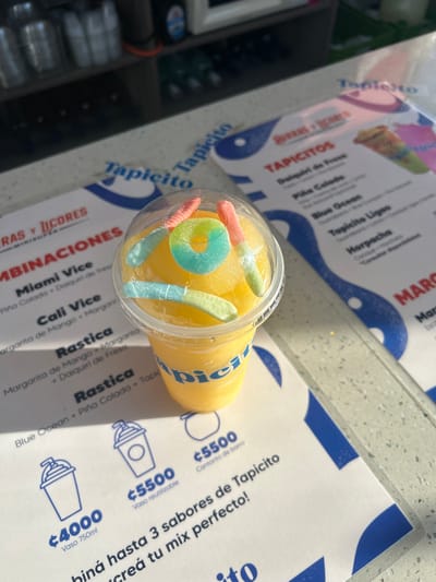 The image shows a clear plastic cup filled with a yellow-colored beverage, topped with gummy candies in various colors, including blue, green, pink, and yellow. The cup is resting on a white, granite-like surface. A blue and white menu sits in front of the cup, featuring the word "Tapicito" printed in bold, blue letters. The menu lists different beverage options, including "Miami Vice," "Cali Vice," and "Rastica." There is also a price list for different sized cups, ranging from €4000 to €5500. The location of the image is likely a restaurant or beverage stand in Pozos, Costa Rica, based on the provided coordinates. The scene is well-lit, suggesting that the image was taken during the day. The overall impression is that of a beverage stand offering a refreshing drink to customers in Pozos.