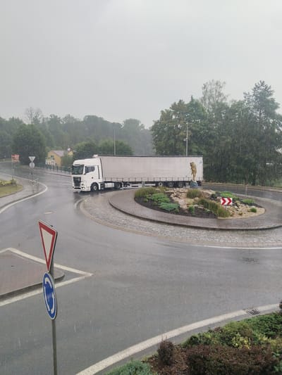 The image captures a white semi-truck driving through a roundabout in what appears to be a semi-urban or rural area. The scene is set in Častolovice, Czechia. It is raining heavily, with visible streaks of rain falling and the road surface appearing wet and reflective. The sky is overcast and grey, suggesting a daytime scene, likely during the day with poor visibility due to the weather.
The roundabout itself is decorated with a circular garden featuring various plants, rocks, and a golden statue of a figure holding a spear or pole. Traffic signs are visible, including a yield sign and a roundabout sign. In the background, there are trees lining the road and a few buildings, including a yellow house. The truck is the main object of focus, as it navigates the curve of the roundabout. The overall mood of the image is subdued, likely due to the heavy rain and overcast conditions. No people are clearly visible in the image, only the truck and the environmental elements.
