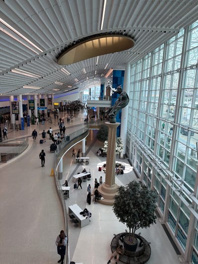 The image shows the interior of a modern airport terminal, likely Charlotte Douglas International Airport in Charlotte, North Carolina, United States of America. The terminal features a high, vaulted ceiling with linear lighting and a large, disc-shaped fixture. The right side of the terminal is dominated by a vast expanse of glass windows, offering a view of the exterior, which appears to include roadways and possibly aircraft.

Prominently featured in the center of the terminal is a large, classical-style sculpture of a draped figure on a pedestal. In front of the sculpture, a tall, artificial tree is planted in a circular planter, surrounded by a curved bench where a person is seated. The floor is a speckled terrazzo material.

The concourse is populated with numerous travelers, many of whom are walking with luggage, seated at tables, or standing in small groups. Several curved railings and platforms delineate different levels and walkways within the terminal. Information kiosks and digital signage are visible in the background, along with a sign indicating "ALL GATES." The overall impression is one of a busy, well-lit, and spacious airport environment.