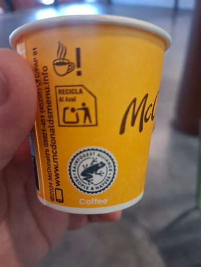 A close-up shot focuses on a yellow McDonald's coffee cup held in a hand. The cup features a white rim and bears several graphics: the McDonald's logo, a recycling symbol indicating "Recicla Al Azul," a Rainforest Alliance Certified seal with a frog, and a cup icon with steam. Text on the cup includes "Coffee," "©2024 McDonald's 03821-451 (45295) C/PAP 81," and "www.mcdonaldsmenu.info." The image suggests the cup is related to a McDonald's location in Spain, and more particularly, to the city of Xirivella, where postal code C/PAP 81 is used. The background is blurred, indicating a shallow depth of field.