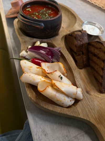 The image shows a wooden platter with various food items. From left to right, there is a dark-colored ceramic pot containing red soup with visible green herbs, next to slices of red onion and garlic cloves, and a small red pepper. Further to the right, there are stacked slices of light-colored meat with a reddish rind, rolled loosely. Above the soup pot is a small silver bowl filled with white sauce, and above that are slices of dark brown bread. The platter sits on a table with a gray surface. The edge of what appears to be a person's denim-clad leg is visible at the bottom right corner of the image. The location is Taraclia, Moldova.