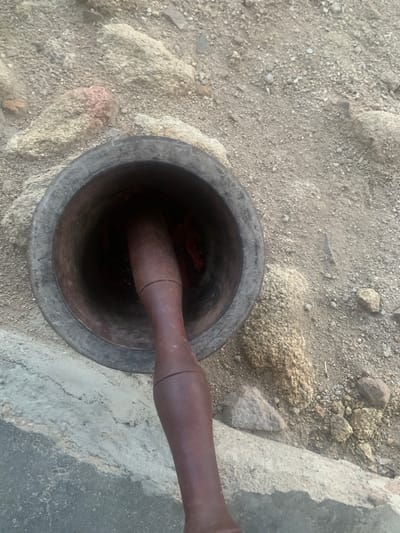 A dark-colored mortar and pestle are positioned on a sandy surface in Gumshir, Nigeria. The mortar is a large, cylindrical stone bowl with a rough texture, and it appears to be partially filled with dark red contents. The pestle, made of wood and a deep brown color, is inserted into the mortar, suggesting that it is in active use or has been recently used. The surrounding ground is composed of sand and small rocks, with larger, light-colored stones scattered around the mortar. The scene is outdoors, and the lighting suggests it is daytime. There are no people visible in the frame. The overall impression is one of a tool used for food preparation in a rustic or natural environment.