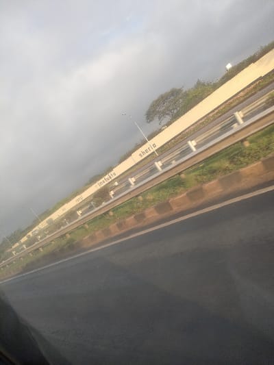 This image presents a diagonally oriented, matter-of-fact view of a multi-lane highway in Theta ward, Kenya, under an overcast sky. The foreground consists of a dark asphalt road surface, appearing slightly reflective. To the right, a brown concrete curb separates the asphalt from a strip of green grass. Beyond this, a silver-colored metal guardrail system runs parallel to the road. Behind the guardrail, another band of green vegetation precedes a tall, light-colored concrete barrier wall.

Prominently displayed on the concrete barrier, painted in black capital letters, are the Swahili words: "sheria," "Imefuta," "Rangi," and "Hii." Spaced along the wall are multiple streetlights with curved arms extending over the carriageway. In the background, indistinct vehicles are visible on an opposing carriageway behind the guardrail and wall. Sparse green trees and vegetation are visible above the concrete barrier, silhouetted against the grey sky. No individuals are discernible within the scene. The overall perspective, including reflections in the lower left, indicates the photograph was taken from inside a moving vehicle.