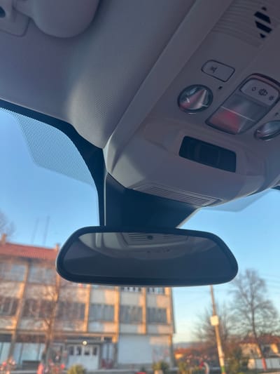 The image provides an interior view from a vehicle, looking forward through the windshield. The upper portion of the frame displays the vehicle's light-colored overhead console, equipped with various buttons, including one with a red indicator, a microphone grill, and a sensor housing. A black-framed rearview mirror is centrally mounted below the console, reflecting elements of the vehicle's interior. Beyond the windshield, a clear blue sky is visible. The external environment, largely out of focus, features two multi-story buildings. The building on the left has a visible reddish-brown tiled roof and a light-toned facade. The building on the right presents a lighter, possibly off-white or light gray, facade with numerous windows. Bare deciduous trees are scattered throughout the background and mid-ground. The scene is illuminated by direct sunlight, casting a warm hue on the buildings. Undefined figures and objects are discernible at street level in the foreground of the external view. This scene is situated in Resilovo, Bulgaria.