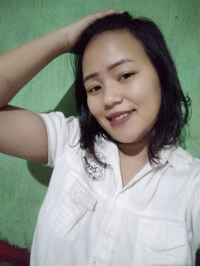 A smiling young woman poses for a selfie. She has short black hair and is wearing a white collared shirt with two front pockets and an embroidered design on the left side. Her left hand is raised to her hair. The background is a green wall. Her expression is cheerful and inviting.