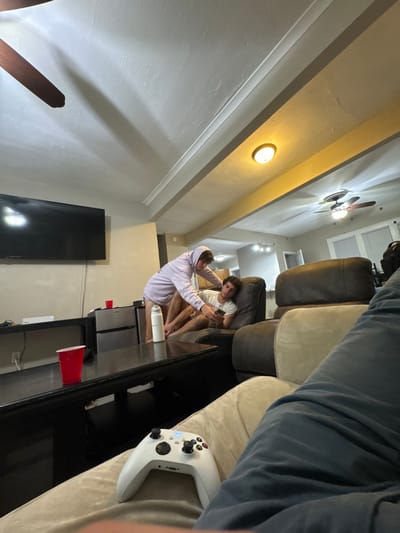 This image depicts an indoor residential living space within South Bend, United States. The scene is captured from a low, seated perspective, showing the observer's legs in dark blue pants with a white Xbox Series X/S controller resting on their lap.
In the midground, a dark wood coffee table features a red plastic cup and a white insulated water bottle. Behind it, a wall-mounted black television is visible above a black media console. To the right of the console, a compact silver and black refrigerator holds a red plastic cup on its top.
Two males occupy the central area. One male, wearing a light purple hooded sweatshirt with the hood up, is standing or leaning over, appearing to interact with a smartphone held by the second male. His lower body is not clearly visible. The second male, wearing a white t-shirt and light-colored shorts, is reclined in a dark brown leather armchair, focused on a smartphone in his hands.
The background reveals an extension of the living space, possibly a kitchen or dining area, with additional lighting fixtures including three track lights and a doorway. The ceiling features light-colored painted surfaces, exposed structural beams with crown molding, and includes two dark-bladed ceiling fans and a circular flush-mount light fixture. The walls are light-colored.