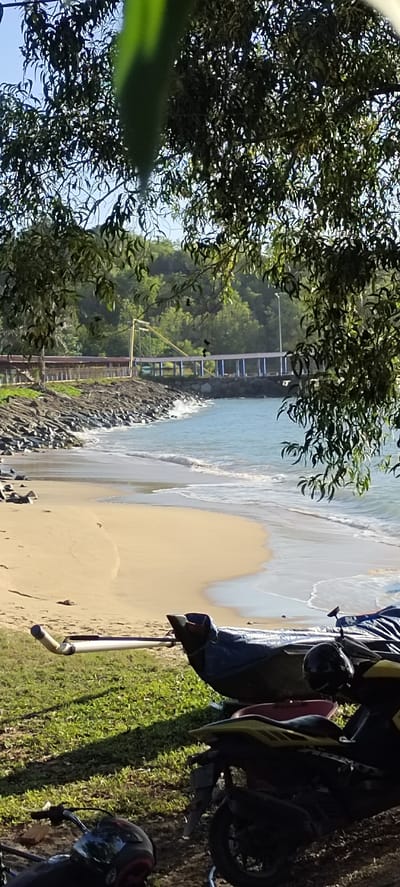 The image depicts a coastal scene in Calang, Indonesia, featuring a sandy beach with gentle waves lapping at the shore. A rocky embankment runs along the edge of the beach, leading to a bridge structure with blue supports and a yellow pylon. Lush green trees frame the top of the image, with their leaves casting shadows. In the foreground, several motorcycles are parked on grassy ground. One motorcycle is yellow and black, with a black helmet resting on its seat. Another motorcycle, partially visible, is darker in color with a red seat. A covered boat or kayak is also present in the foreground, resting on the grass. The sky is clear and blue.