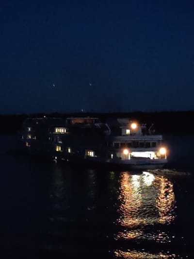 A long, multi-decked passenger ship is illuminated by its own lights as it moves through dark water. The ship is white with several levels, and its windows glow with a warm yellow light. Bright spotlights are also visible on the upper decks. The water reflects the ship's lights in shimmering patterns. The sky is a deep, dark blue with a few faint, blurry lights that may be stars or distant aircraft. The overall impression is a serene, nighttime scene on a body of water, possibly a river or a lake, near the city of Sud'bitsy, Russia.