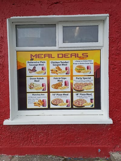 The image shows a bright yellow menu board with various meal deals displayed on a red-painted, textured wall. The menu is behind a window, suggesting it's a takeaway or fast-food establishment. The time of day is not explicitly indicated, but the lighting suggests it could be daytime. There are no people visible in the image.
The menu board features colorful images of food and drinks. The top section prominently displays "MEAL DEALS" in large, stylized letters. Below this, several distinct meal options are presented with accompanying pictures and descriptions. These include:
* **Battered or Plain Sausage Meal:** Depicts sausages and chips.
* **Chicken Tender/Goujon Meal:** Shows chicken tenders and chips.
* **1/4 Burger or Chicken Fillet Burger or Chicken Burger Meal:** Features a burger and chips.
* **Doner Kebab Meal:** Illustrates a doner kebab and chips.
* **Fish & Chips Meal:** Presents a piece of fish with chips.
* **Munchey Box:** Lists the contents including nuggets, tenders, onion rings, chips, doner meat, and sauce.
* **Party Special:** Advertises "Any 5 x 10" Pizza, 3 x Large Chips, Cocktail Sausages & 5 Dips."
* **10" Pizza Meal:** Shows a 10-inch pizza with chips and a drink.
* **Another 10" Pizza Meal:** Displays a 10-inch pizza with chips and a large bottle of Coca-Cola.
Several Coca-Cola branding elements are visible on cans and a bottle, indicating this is likely a fast-food outlet. The overall scene is of a commercial establishment, likely a takeaway food shop, with its menu displayed for customers. The red exterior paint and the style of the window suggest a potentially older building. The location context of Annagry, Ireland, implies a local eatery in a coastal or rural setting.