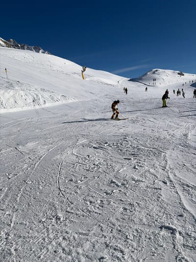 The image displays a snow-covered ski slope under a clear, deep blue sky, located in Klösterle, Austria. The foreground presents a textured snow surface with numerous ski tracks and signs of grooming. Centered on the left, a skier in a dark and yellow-orange suit, wearing a helmet, is actively skiing downhill, creating a slight snow spray from their skis and poles. To the right of this skier, another individual dressed in dark and light green clothing is visible, standing or slowly moving on skis with poles. In the midground and background, approximately fifteen additional individuals, primarily skiers, are dispersed across the expansive white slopes. A yellow snow cannon, mounted on a pole, is situated on the upper left portion of the slope, near a blue and yellow marker pole. Snow-covered mountain peaks are visible in the far background beneath the clear sky, indicating an open ski resort environment.