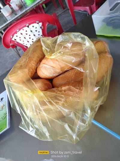 In Gò Dầu, Vietnam, a plastic bag filled with numerous loaves of bread sits on a metal table. The bread, various sizes and shapes, appears to be of the baguette style, with golden-brown crusts. The plastic bag is translucent yellow and obscures some of the bread. In the background, a red plastic chair with a white decorative seat is visible, and a glimpse of a glass table with condiment containers suggests an outdoor or casual dining setting. The scene is brightly lit, casting soft shadows.