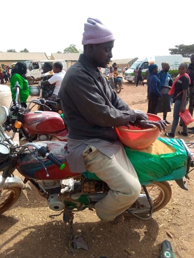 A man in a purple beanie sits on a weathered red motorcycle, holding a red bowl. He wears a gray jacket and light-colored pants. The motorcycle has a green covering over the back. Behind him, people stand around, and several vans and buildings are visible in the background. The ground is brown and dusty.
