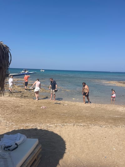 The image depicts a beach scene in Protaras, Cyprus, under a clear blue sky. In the foreground, a thatched umbrella casts a shadow over a white mattress with a bamboo base, with a white shirt draped over it. The sandy beach is strewn with shells. Several people are present. On the left, two young men stand on the shore, one in a white t-shirt and shorts, the other in an orange t-shirt and dark shorts. Further back, a woman in a light pink top and white skirt is looking down, and a man in a striped shirt and khaki shorts stands next to her. In the shallow water to the right, a man in black swim trunks walks, and a young girl in a patterned swimsuit and hat stands, looking at her feet. In the background, a white pier extends into the turquoise sea, with two boats docked and another boat further out. A faint parasail can be seen in the far distance.