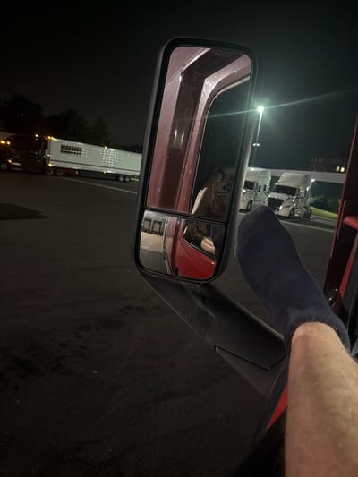 At night, a view from the interior of a semi-truck shows a large side mirror reflecting a person's arm and hand, which is partially covered by a dark blue sock. The arm is extended out of the truck window. In the background, several other semi-trucks are parked. The scene appears to be a truck stop or parking lot. The location is New Britain, Connecticut, United States of America.