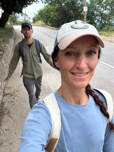 In Malyi Mayak, Ukraine, a man and a woman walk along a paved road beside a stone retaining wall. The man, wearing a dark blue baseball cap and a green long-sleeved shirt over a dark t-shirt, walks with a backpack on. The woman, in the foreground and facing the camera, wears a light-colored baseball cap with a smiley face design and a light blue long-sleeved shirt. She also has a backpack on. The road curves ahead, bordered by trees and vegetation. The weather appears overcast, with some visible dampness on the woman's shirt.