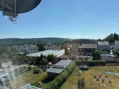The image shows a view of a residential area in Monzelfeld, Germany, on a clear, sunny day. In the foreground, a large satellite dish is visible, partially obscuring the sky. The middle ground features a collection of houses with dark roofs, some with solar panels. A greenhouse with a transparent roof is also present. Beyond the houses, a grassy yard contains a trampoline, a play tent, a fire pit with log seating, and a small blue tarp. In the background, a series of rolling hills covered in dense green forest stretch to the horizon. Several wind turbines are clearly visible on the crests of these hills. The sky is a vibrant, cloudless blue.