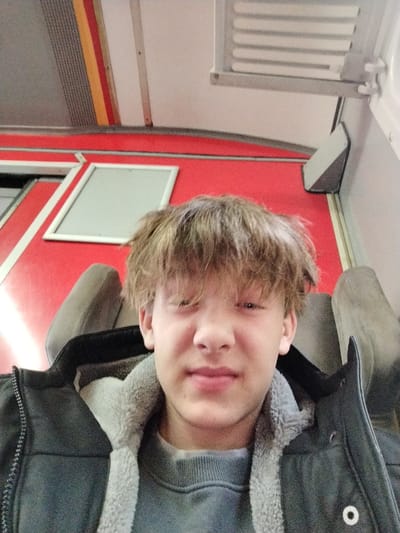 A fair-skinned young man with tousled blonde hair occupies the foreground. He's dressed in a grey sweater and a dark jacket with a grey fleece lining. Behind him, the scene suggests he's on some form of public transport, possibly a train. The interior features red panels, grey seating, and a white ceiling with fixtures. The background colors and architecture point toward an older model transportation design.
