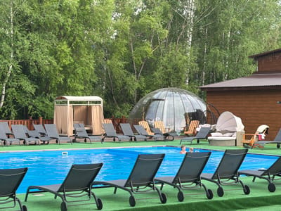 This image captures an outdoor swimming pool area, possibly a resort or spa, situated in a verdant environment. The pool, filled with bright blue water, is surrounded by a vibrant green artificial turf surface. Numerous dark gray lounge chairs are neatly arranged on the turf, facing the pool. In the background, a large, geodesic dome structure with a transparent roof is visible, along with a rustic wooden building. A beige cabana with curtains stands near the pool. The background is dominated by a dense growth of lush green trees. A woman is partially visible in the pool, appearing to swim or relax. The overall scene suggests a recreational area designed for relaxation and enjoyment. The specific location in the city of Tashtagol, Russia is not directly discernible from the visual information provided in the image.
