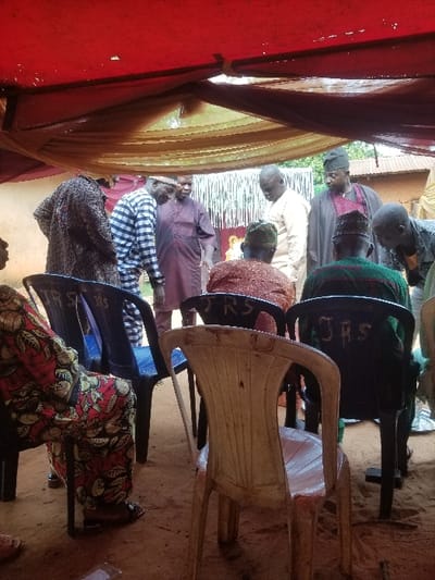 An outdoor gathering is depicted in Ochadam, Nigeria, taking place under a red fabric canopy draped with a lighter, possibly beige, translucent fabric. The ground beneath is bare earth.
In the foreground, a person is seated on the left in a black plastic chair, visible from the mid-thigh down, wearing a brightly patterned garment with red, yellow, black, and green motifs. An empty, light beige plastic chair, showing signs of wear, is positioned centrally with its back to the viewer. Two additional black plastic chairs, each bearing "JRS" in white lettering on the backrest, are partially visible in the mid-foreground, one on the left and one on the right. Two individuals are seated in these chairs, facing away from the viewer; one wears a brownish-red garment and cap, the other a dark green garment and cap.
In the midground, approximately seven men are standing and interacting. From left to right: one man with his back mostly to the viewer, wearing a dark patterned garment; a man in a black and white striped shirt, bending slightly forward and looking down; a man in a dark maroon traditional garment, looking towards the right; a man in a light beige traditional garment, looking downwards; a man in a dark traditional garment and cap, with a visible maroon underlayer, looking towards the left; and a man partially visible on the far right, wearing a light-colored shirt and bending over.
The background reveals a reddish-brown wall structure, possibly mud or brick, with a decorated fabric or wall hanging featuring yellow and orange designs. Above this, corrugated iron roofing is visible, and further behind, green foliage and trees are present. The scene is illuminated by ambient daylight, suggesting an outdoor or semi-outdoor setting.