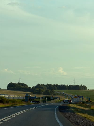 The image depicts a roadside scene in the vicinity of Togashevo, Russia, likely during daylight hours given the illumination. A paved road curves through a landscape of rolling hills and vegetation. Several vehicles, including cars and what appears to be a van, are visible on the road, some moving and others parked. The presence of traffic signs, a bus stop shelter, and a white barrier suggests this is a populated area or a route with infrastructure. In the background, trees form a dense line along the horizon, with power lines and towers indicating the presence of electrical infrastructure. The sky is a pale blue with scattered clouds. The overall impression is of a rural or semi-rural setting on a highway.