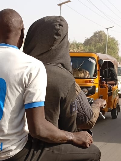 The image depicts an outdoor street scene in Gayawa, Nigeria. In the foreground, two dark-skinned individuals are seated on a vehicle, viewed from behind. The person in front wears a dark grey hooded sweatshirt with the hood up and a "6" insignia on the right sleeve. Scars are visible on their left forearm, and their right hand grips a handlebar, suggesting they are operating the vehicle. The person seated directly behind them has a bald head and wears a white t-shirt with blue sleeve trim. In the midground, a yellow three-wheeled auto-rickshaw (commonly known as a Keke Napep) is partially visible, appearing to be in motion or stopped in traffic. It contains a driver wearing light-colored attire and a passenger in dark clothing. The background includes green foliage, utility poles topped with what appear to be solar street lights, under a bright, clear sky.