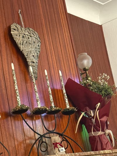The image shows an interior scene, featuring a wall with horizontal wooden paneling. A woven, heart-shaped decoration with a short stem rests against the paneling. Above and to the right of the heart, a decorative candle holder with four arms extends outward. Each arm ends in a flower-patterned rod with a white tip and a small, round, dark-colored decoration at the end. Above the candle holder, there is a wall-mounted lamp with a frosted glass shade. Next to the lamp, a burgundy wrapped bouquet is placed. The overall impression is that of a rustic interior decoration on a wall. The location is not identifiable from the provided visual information.