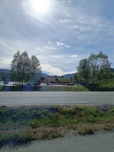 The image shows a bright, sunny day in Hovden, Norway, with a few fluffy white clouds scattered across a light blue sky. The sun is high in the sky, creating a bright glare. In the foreground, a paved road runs horizontally across the frame, with a grassy verge on the right.

Beyond the road, there is a large building with a distinctive yellow roof structure supported by thin pillars. The building appears to be a public facility or a resort, with a red-colored section visible on the left. There are several trees with lush green foliage scattered around the building and the surrounding landscape. To the left of the building, a body of water, possibly a lake or a fjord, is visible, with some picnic tables and outdoor seating areas nearby, suggesting a recreational space. In the background, rolling hills and mountains covered in green vegetation form a scenic backdrop. The overall impression is one of a peaceful, scenic location on a pleasant day, likely during the warmer months. No text is visible in the image.