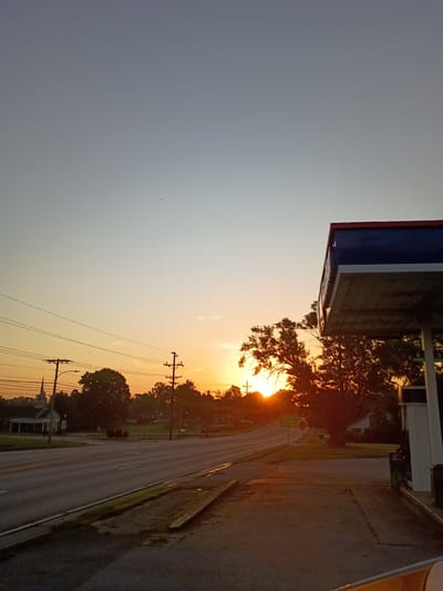 The image captures a roadside scene at sunset in Frankfort, United States of America. The sun, a bright orb, is partially obscured by the branches of a large tree, casting a warm, orange glow across the sky and illuminating the asphalt road. The sky transitions from a soft grey in the upper left to a vibrant sunset orange towards the right.
In the foreground, a portion of a gas station awning with blue and red trim is visible on the left. Adjacent to the road, a sidewalk or paved area can be seen, with some patches of grass and foliage. Telephone poles with electrical wires are prominent features along the side of the road, some with streetlights attached. The road itself appears to be a two-lane highway, with white lines marking the lanes.
In the background, on the right side of the road, are houses and more trees, all silhouetted against the sunset. The overall atmosphere is peaceful and serene, indicative of the end of the day. No people are visible in the frame. There are no discernible signs with text that can be read in this image. The weather appears clear, allowing for an unobstructed sunset view. The time of day is definitively sunset, given the intense colors and the position of the sun.