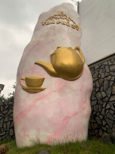 In Thôn 8, Vietnam, a large, pink, rock-like sculpture is prominently displayed. The sculpture features a golden teapot and a golden teacup with a saucer, affixed to its surface. Above these decorative elements, golden letters spell out "TRÀ PHÚ SỸ," likely a brand name or location identifier. The sculpture is situated outdoors, with a stone wall visible on the right side of the image. The ground in front of the sculpture is covered in green grass and some rocks. The sky above is overcast.