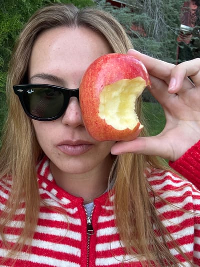 A woman with blonde hair wears black sunglasses and a red and white striped zip-up sweater. She holds a partially eaten red apple over her forehead with her right hand. The image is rotated 90 degrees counterclockwise. Greenery is visible in the background. The woman's lips are painted a light shade of pink. The zipper of her sweater is silver.