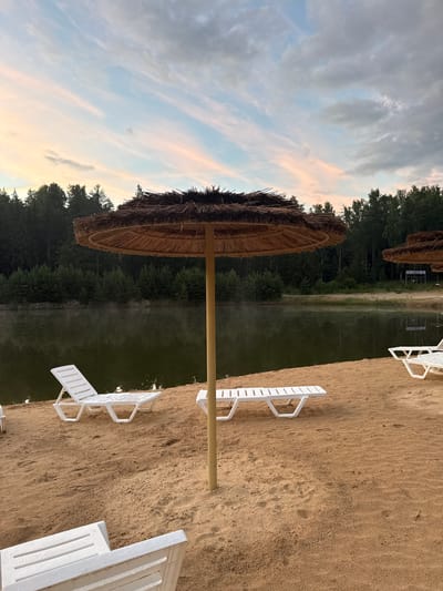 The image depicts a tranquil beach scene at twilight in Sosnovka, Russia. The primary subjects are several empty white plastic lounge chairs arranged on a sandy beach, some partially visible. A large, rustic thatched umbrella stands prominently on the left, its wooden pole emerging from the sand. In the background, a body of calm water, likely a lake or river, is visible, bordered by a dense forest of dark green trees. Wisps of mist or fog are gently rising from the water's surface, adding to the serene atmosphere. The sky above is a canvas of soft twilight colors, with streaks of pale pink and orange blended with soft blues and grays, suggesting the sun has recently set or is about to rise. The overall mood is peaceful and quiet, with no people present, giving the impression of a deserted or early/late hour at a recreational spot. No visible text is present in the image.