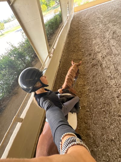 The scene depicts a covered equestrian arena in Rancho Murieta, United States. A person, appearing female, is actively seated on a chestnut-colored horse. The individual wears a black equestrian helmet, a black long-sleeved shirt with a partial button placket, and light grey riding breeches. The left forearm, extended towards the camera for a selfie perspective, displays a silver beaded bracelet and a black and white striped bracelet. The horse is equipped with a dark bridle and a saddle with a white or light-colored saddle pad. The arena floor is composed of light brown, granular equestrian footing, showing numerous hoof prints. To the left, an open side of the arena reveals an outdoor environment featuring green trees, bushes, green grass, and a distant fence line under bright daylight. The arena structure itself consists of large, light-colored support beams and a curved partial wall along the perimeter.