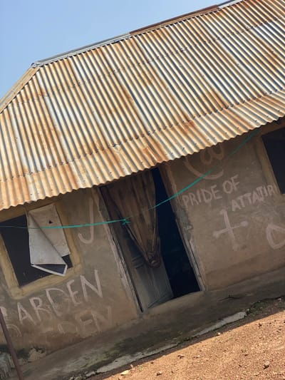 A low-angle, full shot captures a weathered, corrugated metal roofed building constructed of rough concrete. The roof, showing signs of rust and age, slopes downwards from left to right. The walls of the building are a mottled grey, with white paint markings. On the left wall, the word "GARDEN" is painted in large, uneven white letters, with a faded outline of "DEN" below it. A window on the left is covered with a piece of white material and mesh. To the right of this window, a light green string is tied between the wall and a window frame. On the central wall, painted white letters read "PRIDE OF ATTATIRI," with a cross and a circle with a line through it drawn below. A dark doorway is visible in the center of the building, partially obscured by patterned curtains. The ground in front of the building is covered with reddish-brown gravel and small stones. The sky above is clear and blue. The overall impression is one of neglect and decay. The image appears to be taken in a rural or semi-urban setting in Daniel, Nigeria.