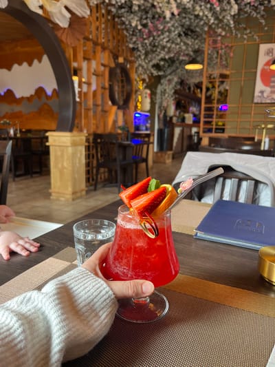 A person holds a red strawberry cocktail adorned with fruit garnishes in a stemmed glass. A clear glass and a baby's hand are visible beside the drink on a brown textured table. In the background, a restaurant interior features a decorative wall, plants, and seating. A blue menu is visible nearby. The scene has a blurred, artistic quality.