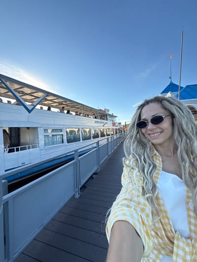 A detailed, forensic description of the scene in Dumino, Russia, shows a bright, clear day at a pier with a passenger boat. In the foreground, a young adult female with light skin and long, blonde dreadlocks/braids smiles, looking slightly past the camera to her right. She wears dark oval sunglasses with silver frames, a white top, and a yellow and white gingham plaid button-up shirt tied at the front. The shirt sleeves are rolled up.
To her left and in the midground, a white passenger riverboat named "МОСКВА-82" (MOSKVA-82/MOSCOW-82) is docked parallel to a dark grey composite or wooden pier. The boat features multiple rectangular windows along its lower deck and an open-air upper deck with a blue and white striped canopy supported by a metal framework. Several individuals are visible on the upper deck. The number "45" is displayed in blue on the boat's hull near the waterline. A light grey metal railing separates the pier from the boat.
In the far midground, behind the woman's head, a building with a distinctive blue roof is partially visible. A flagpole with a small, light-colored flag extends from the roof. The sky above is a clear, bright blue with minimal cloud cover, and sunlight suggests the sun is to the left of the frame.