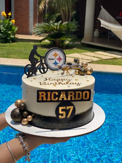 A tilted, eye-level shot presents a birthday cake against the backdrop of a pool. The cake, primarily white with black and gold accents, features the name "Ricardo" and the number "57" in gold lettering. Decorative elements include gold and black spherical adornments, a Steelers logo, and a cutout of a figure on a bicycle. The cake is held by a person wearing bracelets.

The background shows a pool with blue water and surrounding green lawn. A hammock and some vegetation are visible in the distance. The location is identified as San Francisco Lachigoló, Mexico.