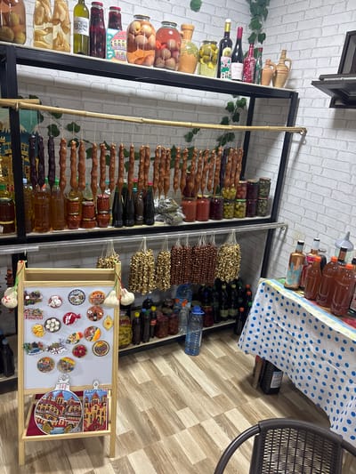 The image displays an indoor market stall or a small shop in Manavi, Georgia, specializing in local products and souvenirs.
The scene features a multi-tiered black metal shelving unit against a white brick-patterned wall. On the top shelf, various glass jars contain preserved fruits (peaches, apples) and possibly olives. Alongside these are bottles, including wine bottles with Georgian labels (e.g., "Saperavi" in Georgian script), and decorative clay pitchers. Artificial green vines are draped on the wall in the upper right quadrant.
The middle shelf features a bamboo rod from which multiple strands of "churchkhela" (grape juice-coated nuts) in dark purple and light brown varieties are suspended. Beneath these, numerous smaller glass jars hold preserves (jams, sauces, pickled items) and various bottles of liquids are arranged.
The bottom shelf holds bundles of dried goods hanging, appearing to be dried fruits and hazelnuts. Below these are numerous plastic and glass bottles containing various liquids.
In the bottom left, a wooden easel-style display stand features a white board adorned with numerous circular and irregularly shaped souvenir magnets. These magnets depict Georgian landmarks (e.g., Tbilisi Old Town), Georgian script, and "TBILISI" in English. On the easel's lower tier, two decorative cutting boards or plaques are displayed. The circular board clearly shows "მანავი" (Manavi) and "საქართველო" (Georgia) in Georgian script, alongside architectural illustrations. The rectangular board also depicts Georgian architecture and "TBILISI" in English.
To the right, a table is covered with a white tablecloth featuring blue polka dots. On this table are several plastic and glass bottles containing amber-colored liquids, and a small funnel is visible behind them. A large black plastic container is partially visible beneath the table.
The floor is covered with light brown wood-grain laminate planks. A dark brown wicker-style chair is partially visible in the bottom right corner of the frame. No individuals are present in the image.
