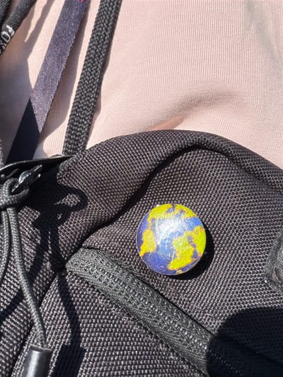 The image shows a close-up of a black backpack. Affixed to the backpack is a small, round object resembling a stylized globe, with green areas representing landmasses and blue areas representing water. The backpack is partially situated on a light tan surface. The presence of a "FOX" logo on a nearby strap suggests it might be a product of Fox Racing. The location of the image is unknown, and there is no indication of it being in Oreshniki, Russia.