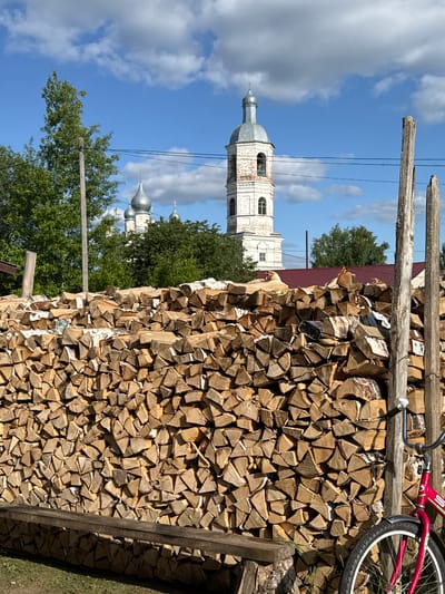 This image depicts a rural or village scene, specified as being in the city of Луптюг, Russia. The foreground is dominated by a meticulously stacked, large pile of split firewood, primarily birch, showing varying shades of light brown and white bark. A rustic, unpainted wooden bench is positioned horizontally at the lower left edge, in front of the firewood. To the right, a partially visible red bicycle with silver fenders and white-walled tires leans against a weathered, rough-hewn wooden post. Another similar wooden post stands further left, behind the woodpile.
Behind the firewood, green deciduous trees obscure the midground. In the background, under a bright blue sky with scattered white cumulus clouds, two distinct church structures are visible. To the right, a tall, white-painted bell tower with multiple arched windows rises, topped by a grey metallic dome and a spire with a cross. To the left of the bell tower, above the treeline, several silver-colored onion domes with crosses are discernible, indicating another church structure. A dark red corrugated metal roof of an unseen building is visible below the bell tower, partially hidden by the woodpile and trees. Thin black power lines traverse the sky horizontally. The scene is illuminated by bright sunlight, casting distinct shadows. No people are present in the image.