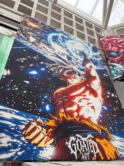 The image shows a large canvas print of the anime character Goku from Dragon Ball Z, in his Ultra Instinct form, depicted in space with the Earth in the background. He is shown powering up with a blue energy aura around him. The canvas is propped up against a white, grid-like structure, possibly a shelf or partition. To the right, another canvas print is partially visible, featuring a pink character with a similar anime art style. The scene appears to be an indoor market or a retail space, possibly a booth at a convention or a shop in El Paso, Texas. The lighting suggests an indoor environment with natural light coming from above, possibly through a skylight or large windows, indicated by the bright white ceiling structure. The canvas prints are the primary focus, showcasing vibrant colors and detailed artwork. The overall impression is that of an art display or merchandise for sale.