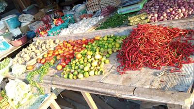 This media file depicts an outdoor market stall in Puloara, Indonesia. The scene is set on a wooden table laden with a variety of fresh produce. The primary subjects are the fruits and vegetables, including a large pile of red chilies, tomatoes, limes, and what appear to be potatoes and garlic. There are also bunches of green chilies and leafy greens. In the background, sacks of what might be rice or flour, bottles of condiments like soy sauce and chili sauce, and bags of snacks are visible. Red onions are piled high in the background.
The activity taking place is the sale of produce at a market. The arrangement of the goods suggests a bustling marketplace where customers can select fresh ingredients. The lighting indicates it is daytime, with sunlight casting shadows across the wooden surfaces and the produce, suggesting a sunny day. The setting is rustic, with the rough-hewn wooden table and the variety of goods giving a sense of a traditional Indonesian market. No people are clearly visible in the frame, but their presence is implied by the market setting.
Notable details include the vibrant colors of the produce, particularly the red chilies and tomatoes. The textures of the different items, from the smooth skins of the limes to the bumpy surfaces of the potatoes, are also discernible. Some text is visible on the packaging of the snacks in the background, including "ABC" logos, suggesting common Indonesian brands. The overall impression is one of abundance and freshness, characteristic of a local food market.