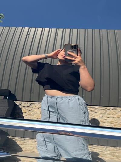 A person is taking a mirror selfie outdoors, standing in front of a corrugated metal wall. They are wearing a black cropped t-shirt and light blue cargo shorts. The person has their right arm raised, touching their head, and is holding a smartphone in their left hand. A silver watch is visible on their left wrist, and a ring is on their left index finger. The background consists of the metal wall, a stone wall below it, and a clear blue sky above. The reflection in a car door in the foreground shows the person's lower body and the surrounding environment. The image appears to have been taken during daylight hours. There is no specific indication of the location being in Velyki Hai, Ukraine.