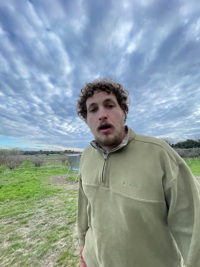 The image displays a male subject in a rural, agricultural environment in Cucuron, France. The man, positioned centrally in the foreground, has curly brown hair and a light beard, looking directly at the camera with his mouth slightly agape. He wears an olive-green Columbia quarter-zip fleece pullover. A small red object or fabric is visible near his lower left side. Behind him, to the left, is a silver-toned, open-top metal utility cart on wheels. The background consists of a field with green grass and visible dirt paths, lined with rows of what appear to be dormant fruit trees or bushes. The sky is largely covered by a pattern of alto-cumulus clouds.