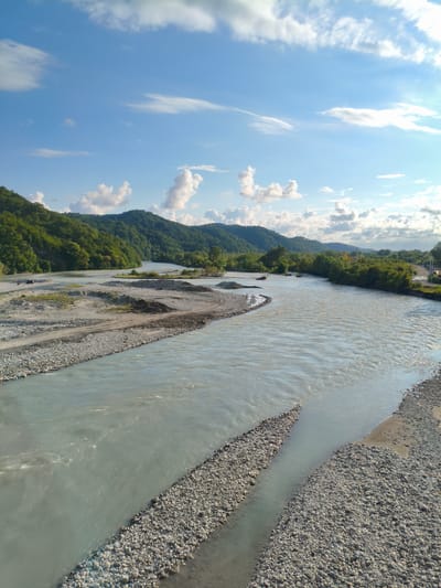 The image captures a wide, slow-moving river flowing through a valley. The riverbed is lined with grey and white pebbles, with some areas of sand and sediment visible. Lush green trees cover the rolling hills that flank the river on both sides. The sky above is a bright blue, dotted with fluffy white clouds, suggesting a pleasant day.

The scene appears to be a natural landscape, possibly in a rural or mountainous region. The presence of a road and some structures in the distance, particularly on the right side of the image, hints at some human presence or development in the area, but the overall impression is one of natural beauty. There are no people or specific objects that draw the viewer's attention, allowing the focus to remain on the grandeur of the landscape. The time of day appears to be either late morning or early afternoon, given the bright sunlight and the shadows cast. The weather seems fair and clear. The location context provided, Tsaniguarta, Abkhazia, places this scene within a specific geographical area known for its scenic beauty. The image conveys a sense of tranquility and the raw power of nature.