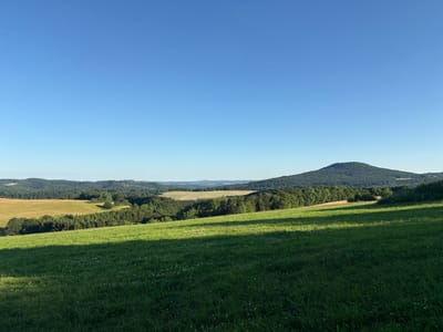 The image depicts a serene landscape in Wershofen, Germany, characterized by rolling green hills under a clear blue sky. In the foreground, a lush green meadow slopes gently downward, dotted with small white wildflowers. A distinct shadow falls across the meadow from the left, suggesting the presence of an unseen object or obstruction casting the shade.

The middle ground is a patchwork of agricultural fields and dense forests. A field of golden-brown crops is visible, bordered by a line of dark green coniferous trees. Beyond this, a valley opens up, revealing more forested hills that stretch towards the horizon.

Dominating the right side of the background is a prominent, conical hill densely covered in trees. The hill's silhouette is sharp against the bright sky, indicating it is a significant geographical feature in the area. The overall atmosphere is tranquil and natural, with no visible human presence or activity, emphasizing the rural and picturesque setting of Wershofen. The image appears to have been taken during daylight hours, likely in the late afternoon due to the long shadows.