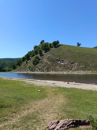 The image shows a landscape scene. In the foreground, there is a green meadow with some rocks scattered around. A body of water, possibly a river, stretches across the middle ground. The water is calm and reflects the sky. Beyond the water, there is a hill covered in green grass and dotted with trees. The hillside appears to have some rocky outcrops as well. In the background, there is a forested area and a clear blue sky above. Gabdyukovo, Russia is not explicitly identifiable from the imagery.
