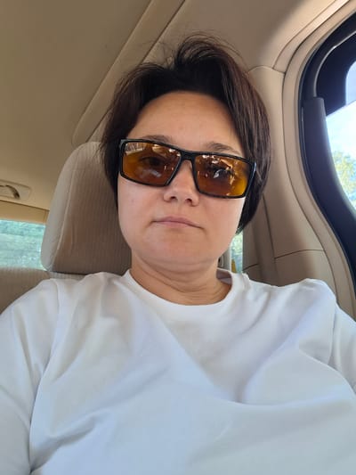 The image is a close-up, indoor selfie of a person in a car. The subject, a woman with short dark hair and wearing sunglasses, is looking directly at the camera. She is wearing a plain white t-shirt. The background shows the interior of a vehicle, with a beige headrest and car door visible. The lighting suggests it is daytime. The location is identified as Svobodny, Russia.